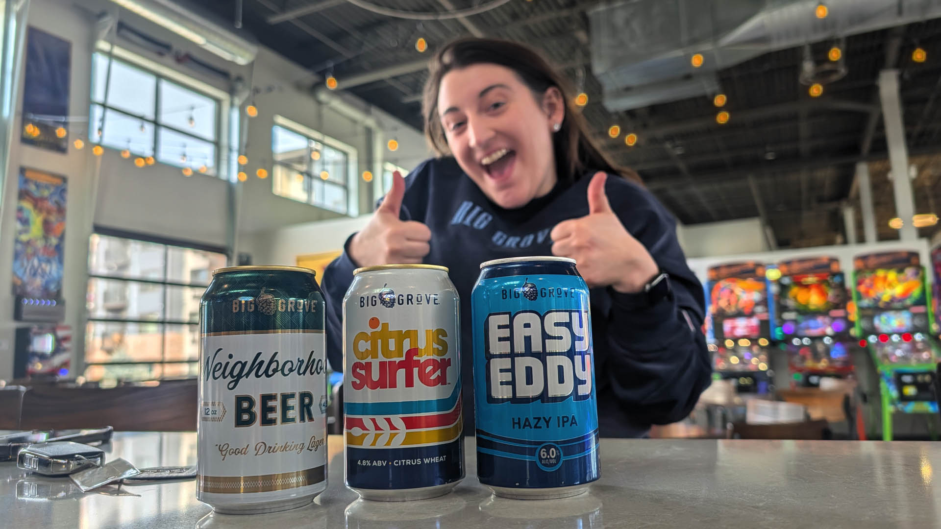 Three Big Grove beers on a bar with someone giving double thumbs up behind them.